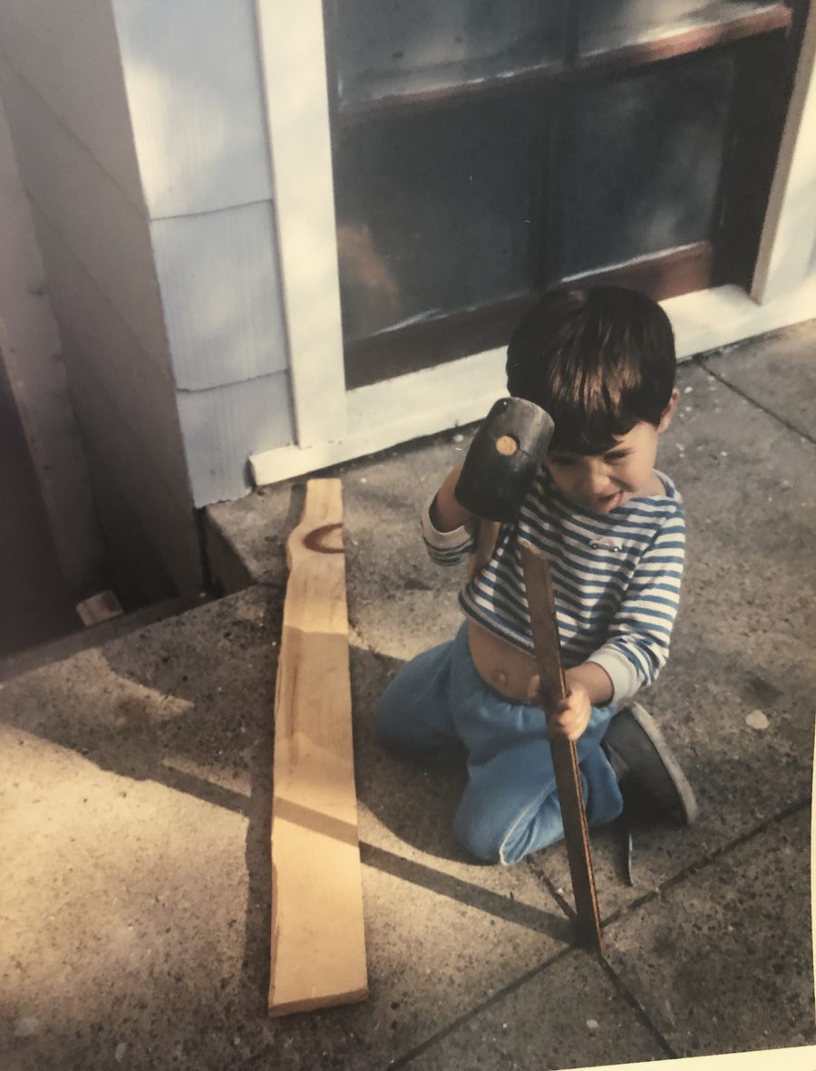 Preschool-age me kneeling on the sidewalk, grinning, with a mallet raised over a wooden stake.